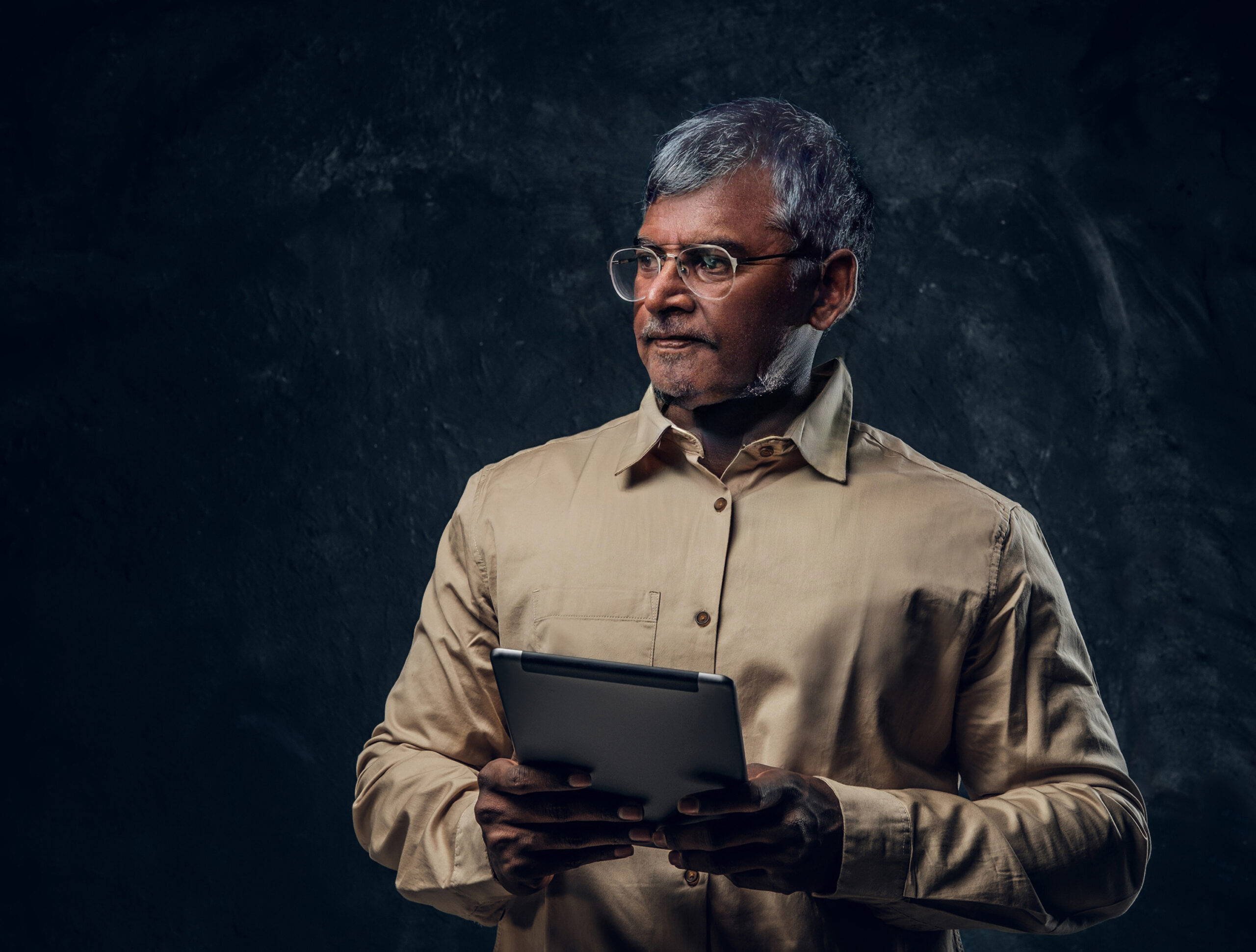 Studio shot of hindu grandfather dressed in beige shirt using tablet against dark background.