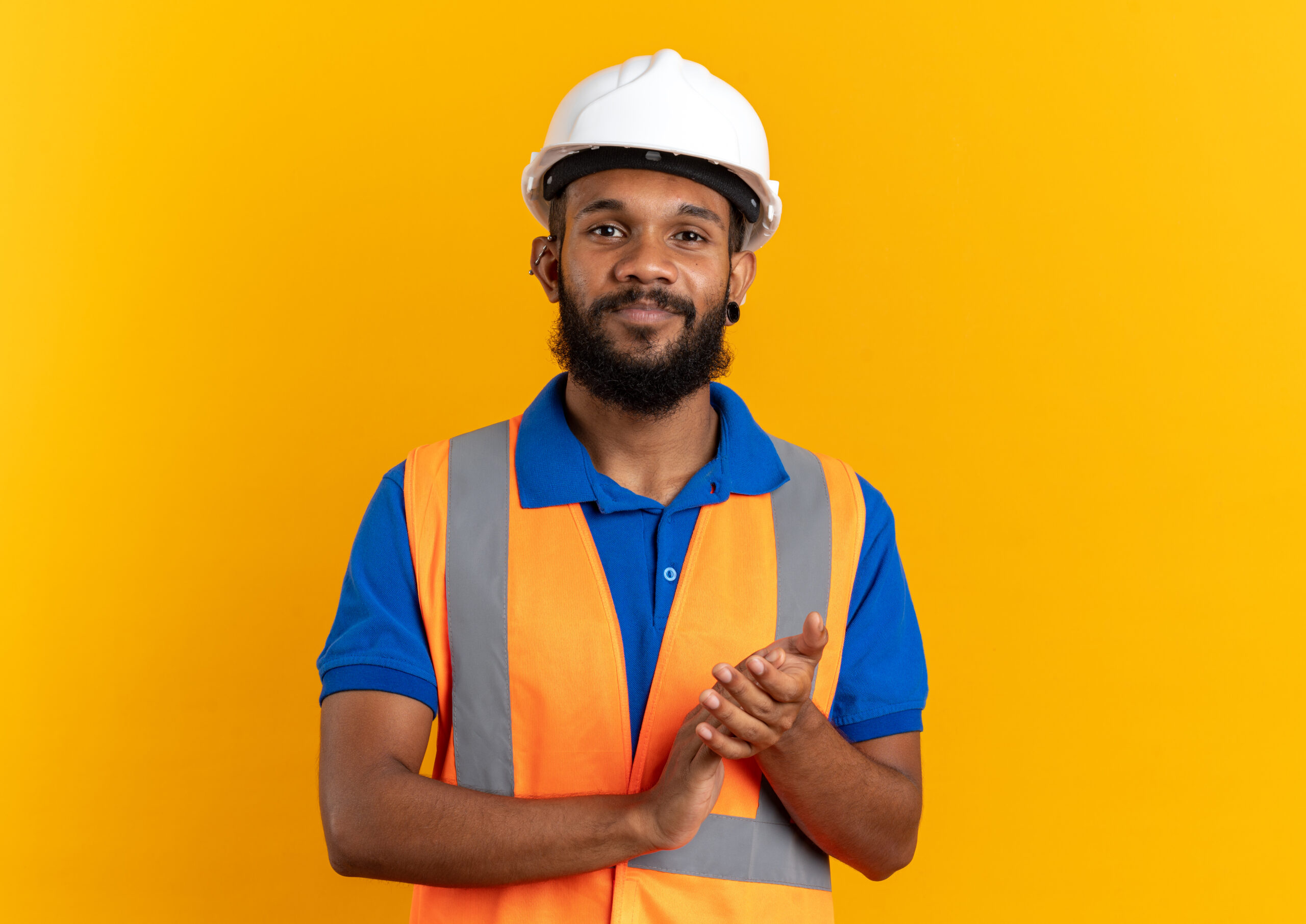 pleased young afro-american builder man in uniform with safety helmet holding hands together isolated on orange background with copy space